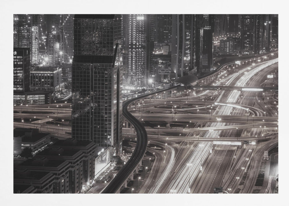 A dynamic black and white long-exposure photograph of a sprawling metropolis at night, viewed from above. Gleaming skyscrapers fill the background, while a complex interchange of highways with streaks of light from traffic dominates the foreground, with an elevated train line weaving through the city. Wall Art