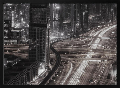 A dynamic black and white long-exposure photograph of a sprawling metropolis at night, viewed from above. Gleaming skyscrapers fill the background, while a complex interchange of highways with streaks of light from traffic dominates the foreground, with an elevated train line weaving through the city. Wall Art