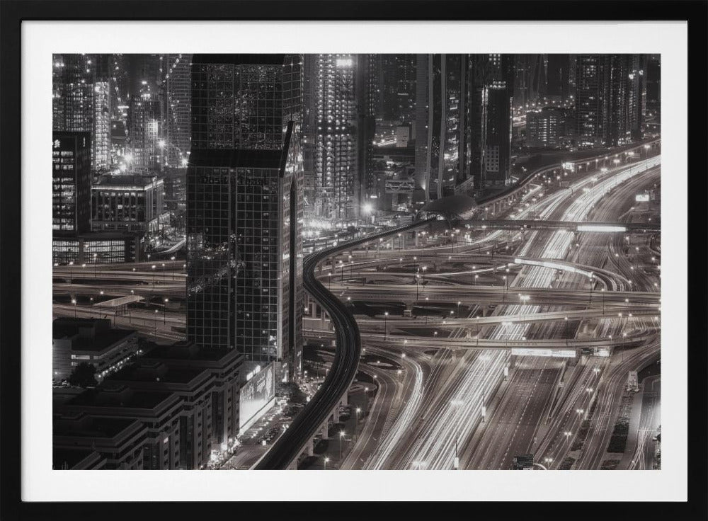 A dynamic black and white long-exposure photograph of a sprawling metropolis at night, viewed from above. Gleaming skyscrapers fill the background, while a complex interchange of highways with streaks of light from traffic dominates the foreground, with an elevated train line weaving through the city. Wall Art