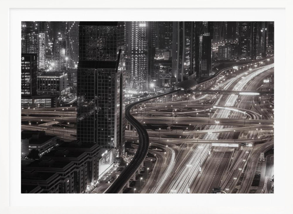 A dynamic black and white long-exposure photograph of a sprawling metropolis at night, viewed from above. Gleaming skyscrapers fill the background, while a complex interchange of highways with streaks of light from traffic dominates the foreground, with an elevated train line weaving through the city. Wall Art