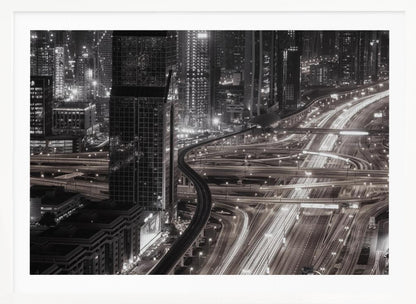 A dynamic black and white long-exposure photograph of a sprawling metropolis at night, viewed from above. Gleaming skyscrapers fill the background, while a complex interchange of highways with streaks of light from traffic dominates the foreground, with an elevated train line weaving through the city. Wall Art