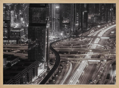 A dynamic black and white long-exposure photograph of a sprawling metropolis at night, viewed from above. Gleaming skyscrapers fill the background, while a complex interchange of highways with streaks of light from traffic dominates the foreground, with an elevated train line weaving through the city. Wall Art