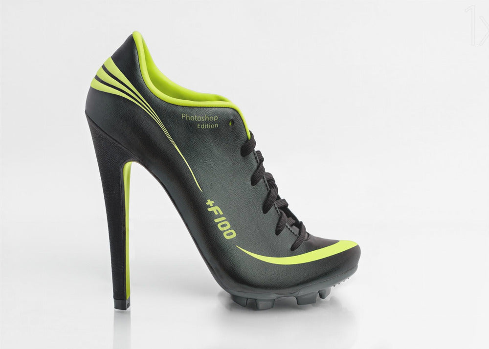 A conceptual piece of digital art featuring a hybrid shoe that is part black soccer cleat and part stiletto high heel. The shoe has neon lime green accents and is displayed against a stark white background inside a silver frame. Decor