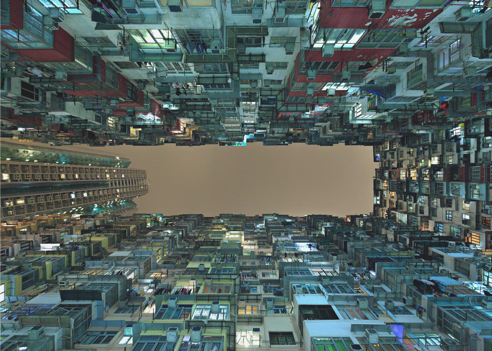 A framed, low-angle photograph looking up from a courtyard at densely packed, colorful apartment buildings that form a rectangle around a blank, overcast sky at night. Print