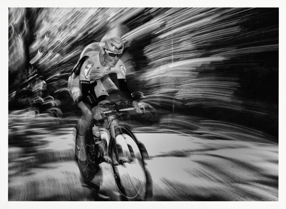 A dramatic black and white action shot of a professional cyclist racing. The cyclist is in focus, wearing a helmet and sunglasses, while the background is a streaky motion blur, creating a powerful sense of speed and movement. The image is enclosed in a silver frame. Wall Art