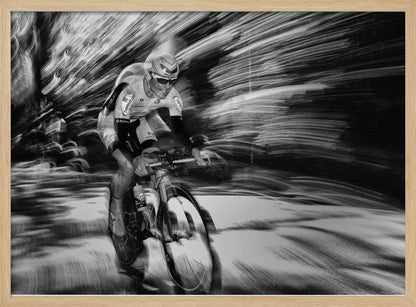 A dramatic black and white action shot of a professional cyclist racing. The cyclist is in focus, wearing a helmet and sunglasses, while the background is a streaky motion blur, creating a powerful sense of speed and movement. The image is enclosed in a silver frame. Wall Art