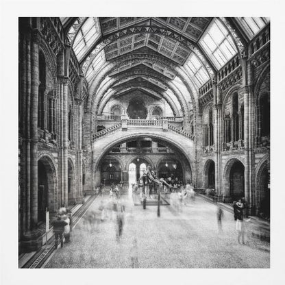 A black and white long exposure photograph of the grand hall of a natural history museum. The ornate, high-arched Romanesque architecture is in sharp focus, while a dinosaur skeleton stands in the center. Crowds of people are blurred into ghostly apparitions as they move through the vast space. Print