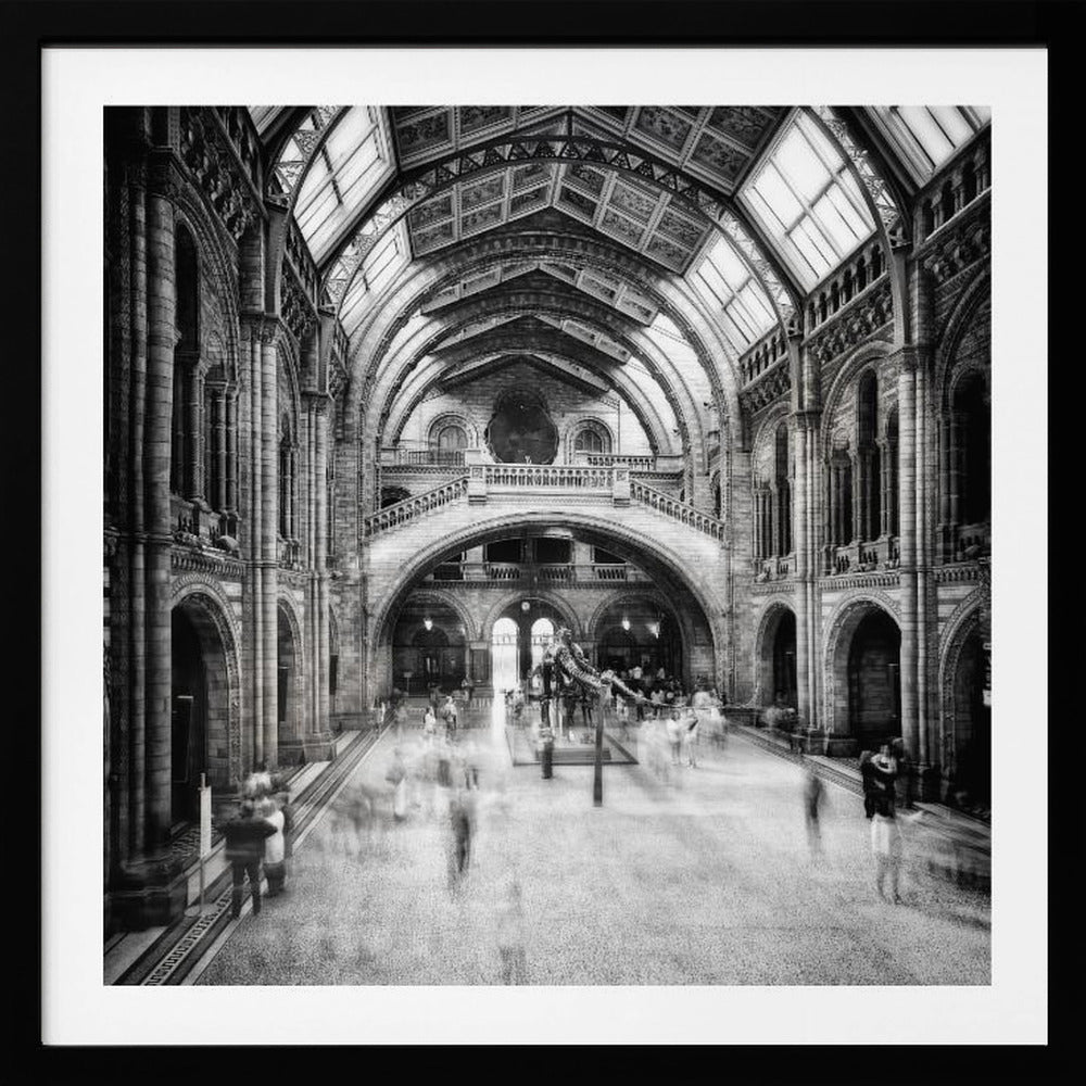 A black and white long exposure photograph of the grand hall of a natural history museum. The ornate, high-arched Romanesque architecture is in sharp focus, while a dinosaur skeleton stands in the center. Crowds of people are blurred into ghostly apparitions as they move through the vast space. Print