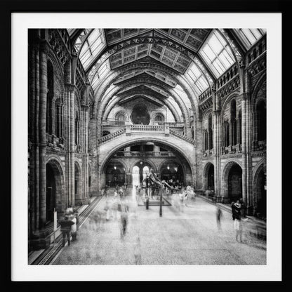 A black and white long exposure photograph of the grand hall of a natural history museum. The ornate, high-arched Romanesque architecture is in sharp focus, while a dinosaur skeleton stands in the center. Crowds of people are blurred into ghostly apparitions as they move through the vast space. Print