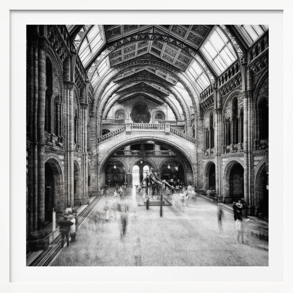 A black and white long exposure photograph of the grand hall of a natural history museum. The ornate, high-arched Romanesque architecture is in sharp focus, while a dinosaur skeleton stands in the center. Crowds of people are blurred into ghostly apparitions as they move through the vast space. Print