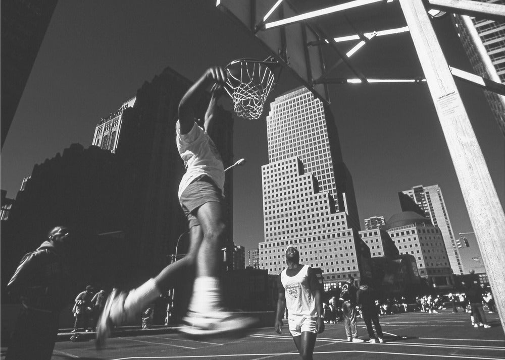 A low-angle, black and white action shot of a person dunking a basketball on an outdoor court, with tall city skyscrapers dominating the background under a clear sky. Print