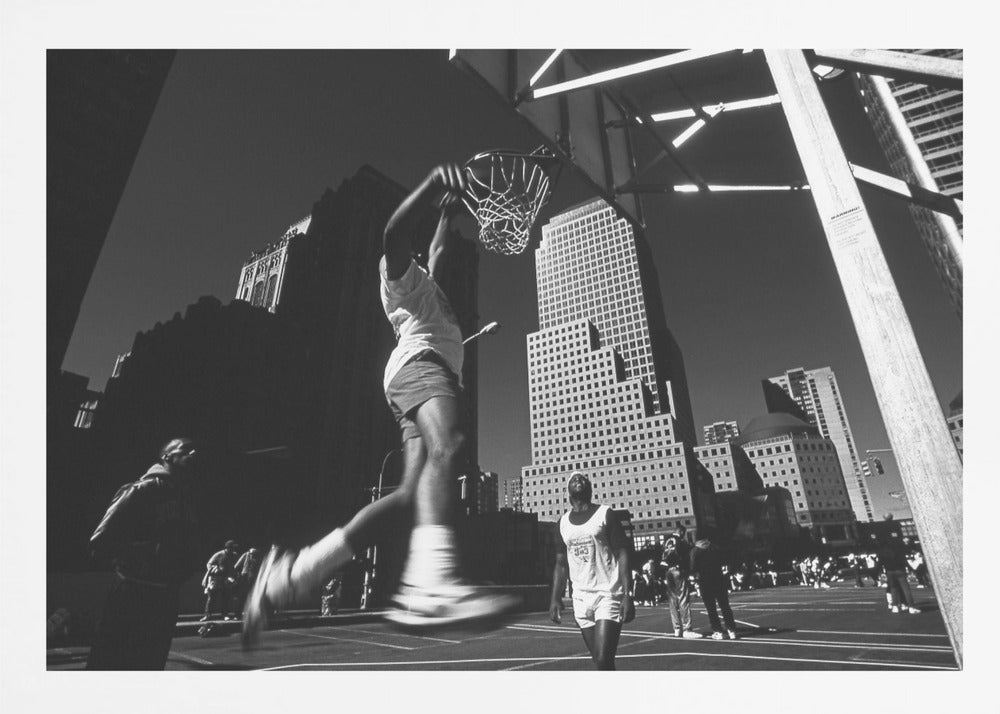 A low-angle, black and white action shot of a person dunking a basketball on an outdoor court, with tall city skyscrapers dominating the background under a clear sky. Print