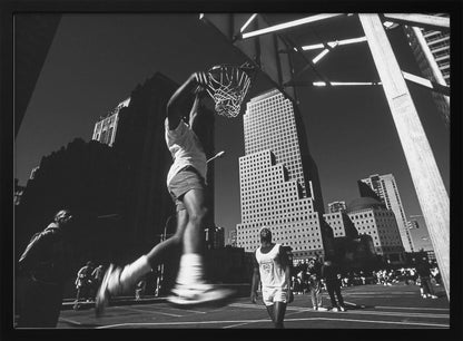 A low-angle, black and white action shot of a person dunking a basketball on an outdoor court, with tall city skyscrapers dominating the background under a clear sky. Print
