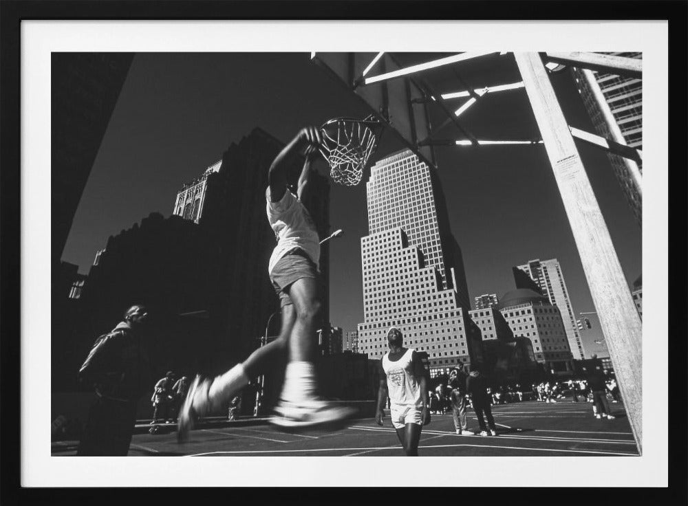 A low-angle, black and white action shot of a person dunking a basketball on an outdoor court, with tall city skyscrapers dominating the background under a clear sky. Print