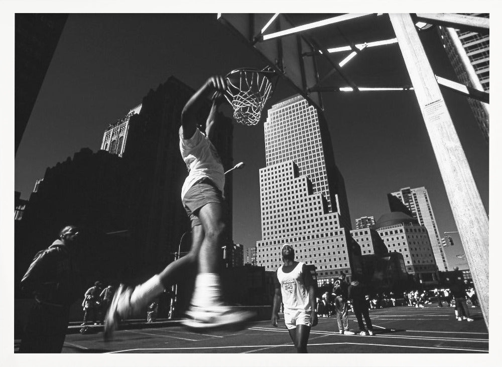 A low-angle, black and white action shot of a person dunking a basketball on an outdoor court, with tall city skyscrapers dominating the background under a clear sky. Print