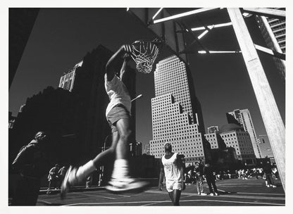 A low-angle, black and white action shot of a person dunking a basketball on an outdoor court, with tall city skyscrapers dominating the background under a clear sky. Print