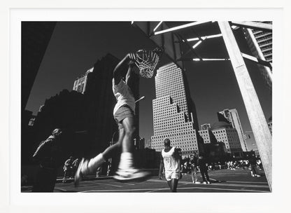 A low-angle, black and white action shot of a person dunking a basketball on an outdoor court, with tall city skyscrapers dominating the background under a clear sky. Print