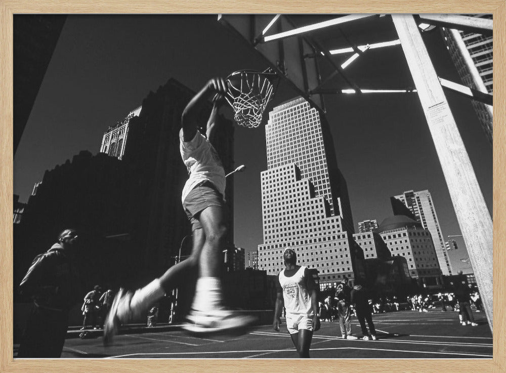 A low-angle, black and white action shot of a person dunking a basketball on an outdoor court, with tall city skyscrapers dominating the background under a clear sky. Print