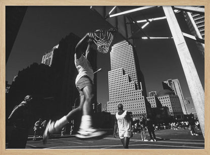 A low-angle, black and white action shot of a person dunking a basketball on an outdoor court, with tall city skyscrapers dominating the background under a clear sky. Print
