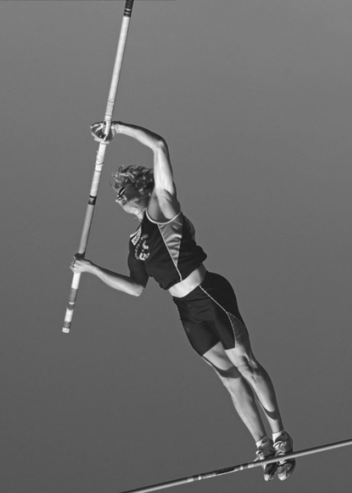 A dynamic black and white, low-angle shot of a female pole vaulter in mid-action. She is suspended in the air, gripping the pole as she prepares to clear the high bar, with her muscular form silhouetted against a plain sky. Print