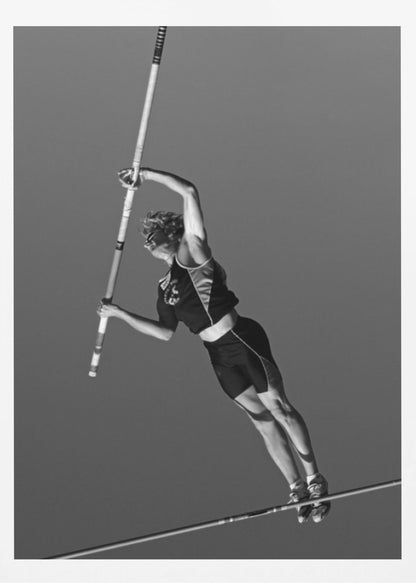 A dynamic black and white, low-angle shot of a female pole vaulter in mid-action. She is suspended in the air, gripping the pole as she prepares to clear the high bar, with her muscular form silhouetted against a plain sky. Print