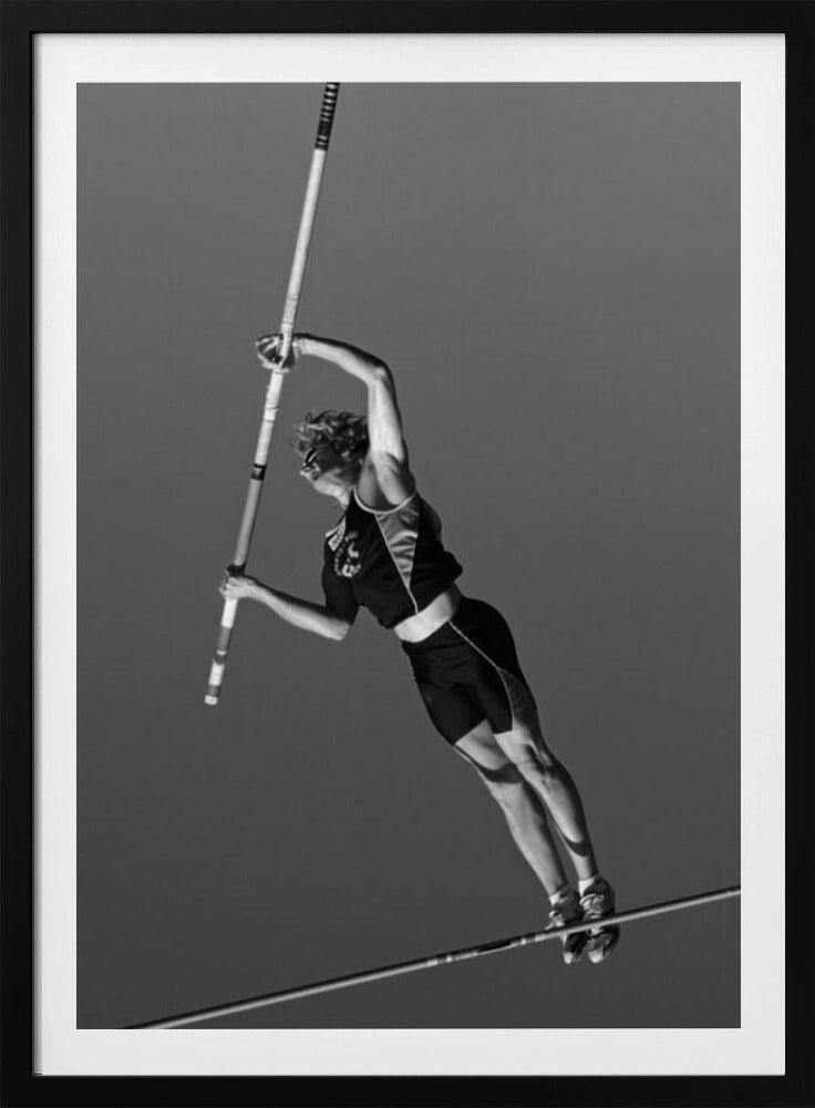 A dynamic black and white, low-angle shot of a female pole vaulter in mid-action. She is suspended in the air, gripping the pole as she prepares to clear the high bar, with her muscular form silhouetted against a plain sky. Print