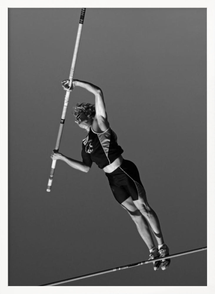 A dynamic black and white, low-angle shot of a female pole vaulter in mid-action. She is suspended in the air, gripping the pole as she prepares to clear the high bar, with her muscular form silhouetted against a plain sky. Print