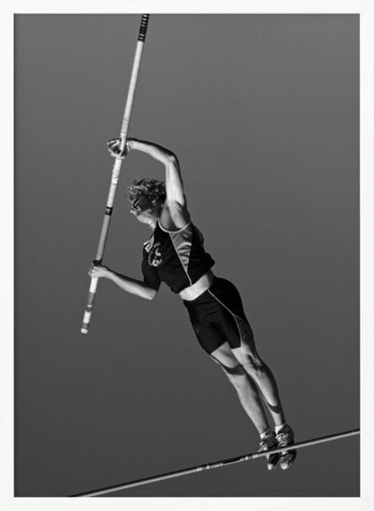 A dynamic black and white, low-angle shot of a female pole vaulter in mid-action. She is suspended in the air, gripping the pole as she prepares to clear the high bar, with her muscular form silhouetted against a plain sky. Print