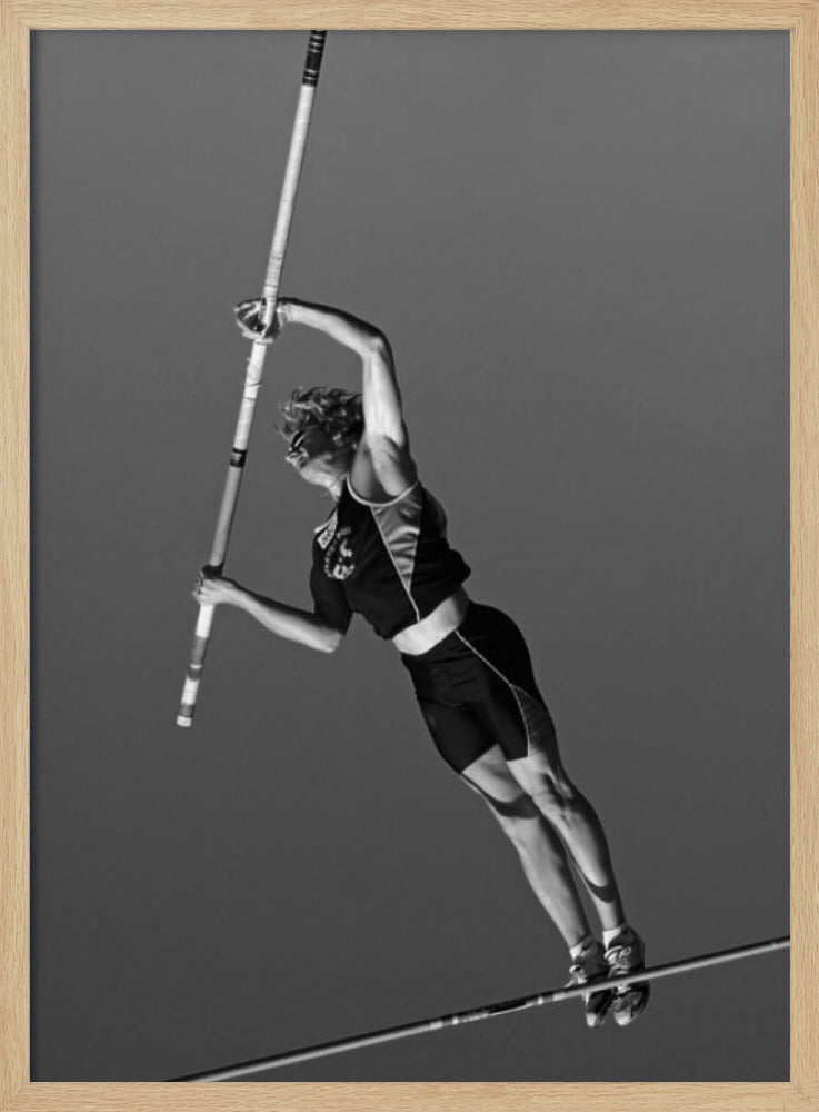 A dynamic black and white, low-angle shot of a female pole vaulter in mid-action. She is suspended in the air, gripping the pole as she prepares to clear the high bar, with her muscular form silhouetted against a plain sky. Print