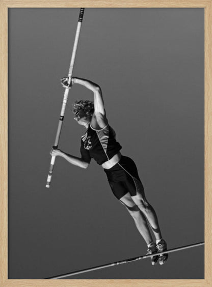 A dynamic black and white, low-angle shot of a female pole vaulter in mid-action. She is suspended in the air, gripping the pole as she prepares to clear the high bar, with her muscular form silhouetted against a plain sky. Print