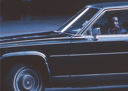 A moody, blue-toned photograph of a Black man sitting pensively in the driver's seat of a dark vintage sedan at night, framed in silver. Poster
