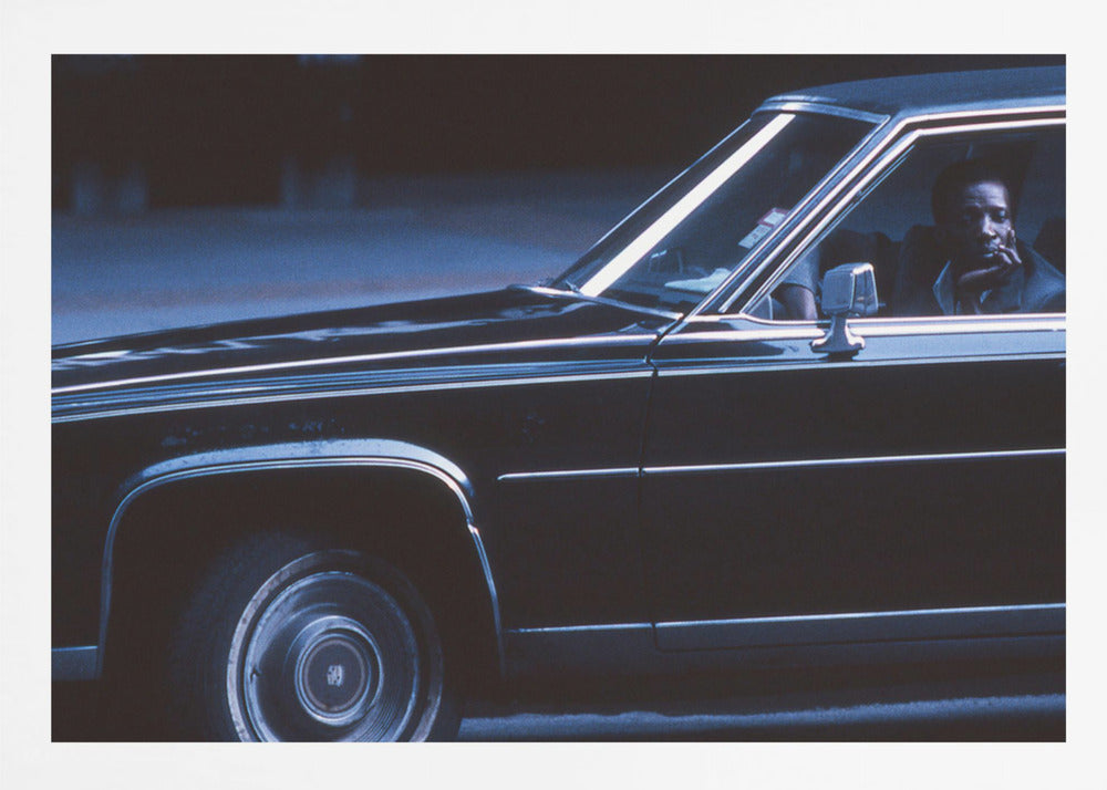 A moody, blue-toned photograph of a Black man sitting pensively in the driver's seat of a dark vintage sedan at night, framed in silver. Poster