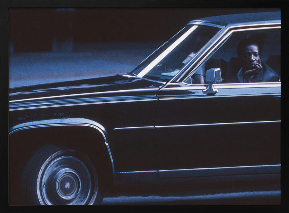 A moody, blue-toned photograph of a Black man sitting pensively in the driver's seat of a dark vintage sedan at night, framed in silver. Poster