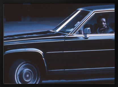 A moody, blue-toned photograph of a Black man sitting pensively in the driver's seat of a dark vintage sedan at night, framed in silver. Poster
