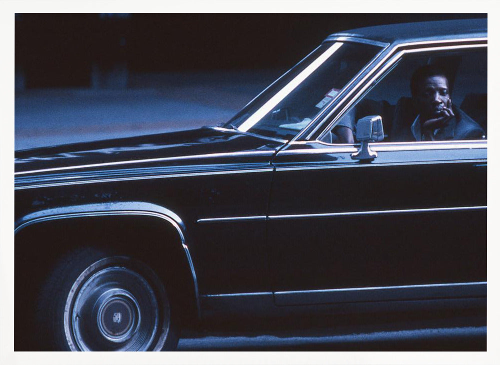 A moody, blue-toned photograph of a Black man sitting pensively in the driver's seat of a dark vintage sedan at night, framed in silver. Poster
