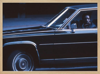 A moody, blue-toned photograph of a Black man sitting pensively in the driver's seat of a dark vintage sedan at night, framed in silver. Poster