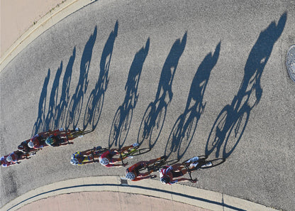 An aerial, high-angle photograph of a line of cyclists racing on a curved asphalt road. The bright sun casts long, dark, dramatic shadows of the riders and their bicycles, creating a striking diagonal pattern across the pavement. The image is framed in silver. Print