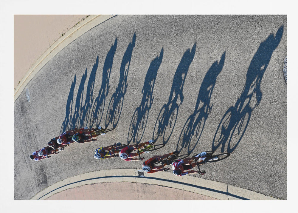An aerial, high-angle photograph of a line of cyclists racing on a curved asphalt road. The bright sun casts long, dark, dramatic shadows of the riders and their bicycles, creating a striking diagonal pattern across the pavement. The image is framed in silver. Print