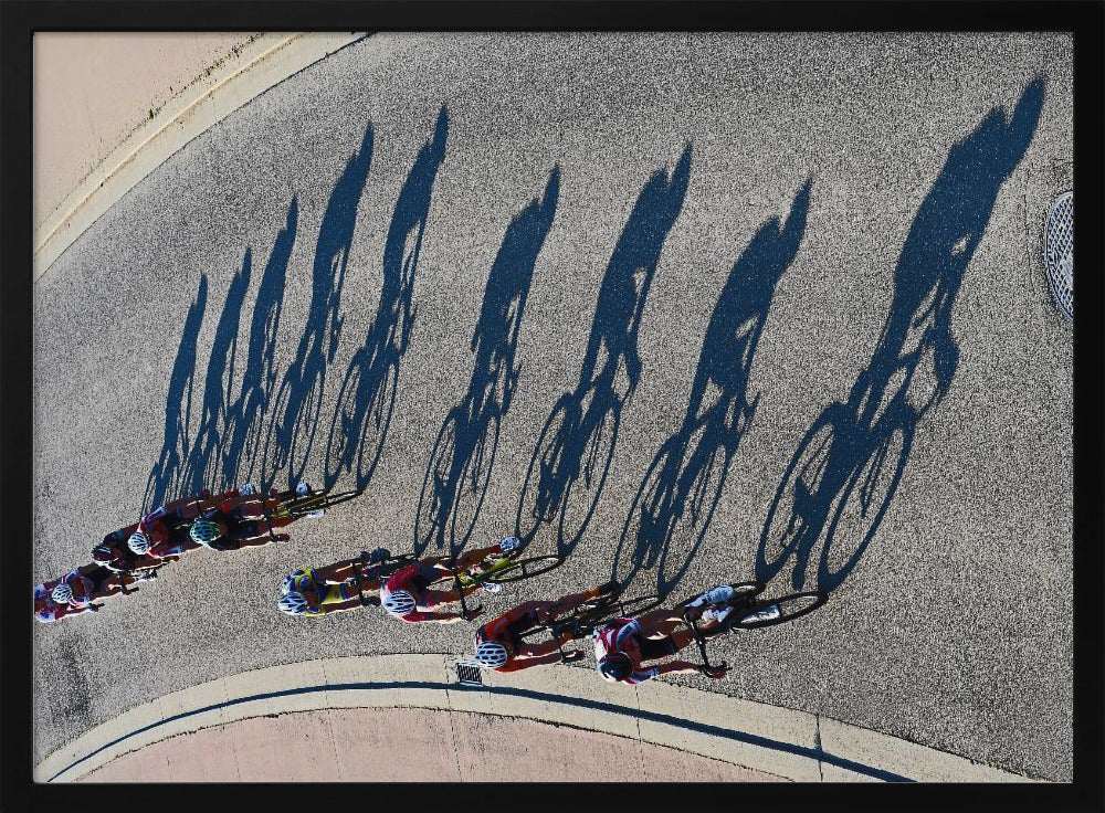 An aerial, high-angle photograph of a line of cyclists racing on a curved asphalt road. The bright sun casts long, dark, dramatic shadows of the riders and their bicycles, creating a striking diagonal pattern across the pavement. The image is framed in silver. Print