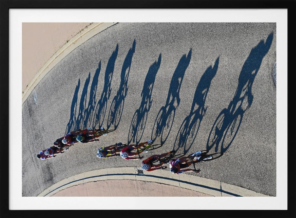 An aerial, high-angle photograph of a line of cyclists racing on a curved asphalt road. The bright sun casts long, dark, dramatic shadows of the riders and their bicycles, creating a striking diagonal pattern across the pavement. The image is framed in silver. Print