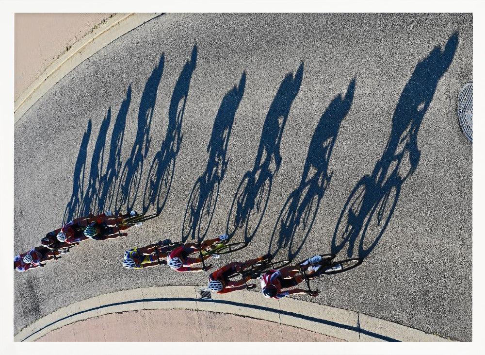 An aerial, high-angle photograph of a line of cyclists racing on a curved asphalt road. The bright sun casts long, dark, dramatic shadows of the riders and their bicycles, creating a striking diagonal pattern across the pavement. The image is framed in silver. Print