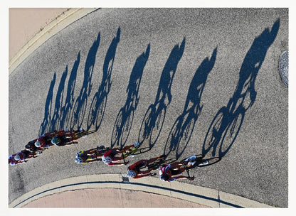 An aerial, high-angle photograph of a line of cyclists racing on a curved asphalt road. The bright sun casts long, dark, dramatic shadows of the riders and their bicycles, creating a striking diagonal pattern across the pavement. The image is framed in silver. Print