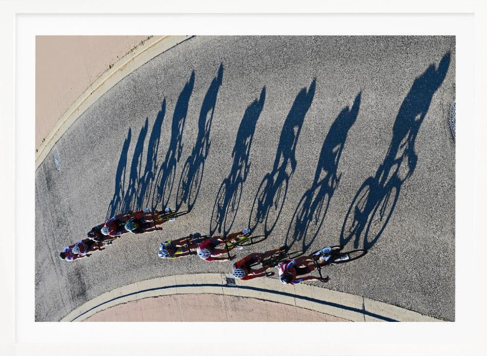 An aerial, high-angle photograph of a line of cyclists racing on a curved asphalt road. The bright sun casts long, dark, dramatic shadows of the riders and their bicycles, creating a striking diagonal pattern across the pavement. The image is framed in silver. Print