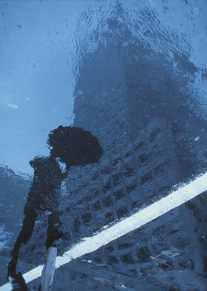 A moody, blue-toned photograph of a reflection in a rainy puddle. A black silhouette of a person holding an umbrella and a distorted brick building are visible, with a single white line crossing the wet street. Poster