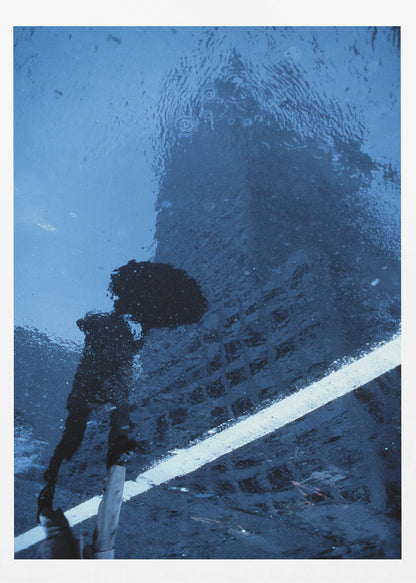 A moody, blue-toned photograph of a reflection in a rainy puddle. A black silhouette of a person holding an umbrella and a distorted brick building are visible, with a single white line crossing the wet street. Poster