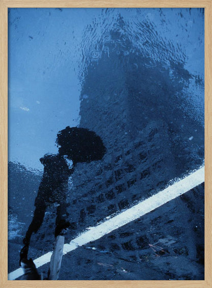 A moody, blue-toned photograph of a reflection in a rainy puddle. A black silhouette of a person holding an umbrella and a distorted brick building are visible, with a single white line crossing the wet street. Poster