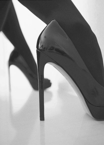 A close-up, black and white photograph of a person wearing shiny black patent leather stilettos and dark stockings. One shoe is in sharp focus in the foreground, highlighting its high arch and thin heel, while the other leg and shoe are softly blurred in the background on a reflective white surface. Decor