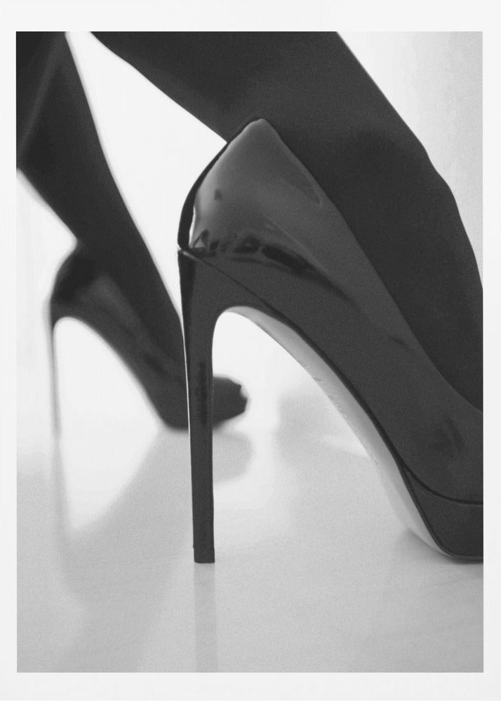 A close-up, black and white photograph of a person wearing shiny black patent leather stilettos and dark stockings. One shoe is in sharp focus in the foreground, highlighting its high arch and thin heel, while the other leg and shoe are softly blurred in the background on a reflective white surface. Decor