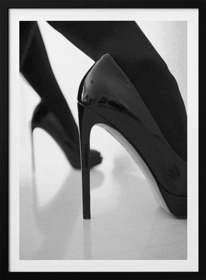 A close-up, black and white photograph of a person wearing shiny black patent leather stilettos and dark stockings. One shoe is in sharp focus in the foreground, highlighting its high arch and thin heel, while the other leg and shoe are softly blurred in the background on a reflective white surface. Decor