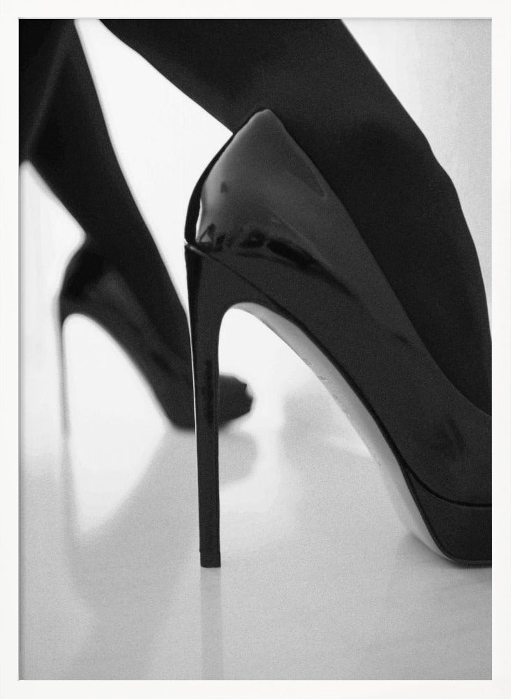 A close-up, black and white photograph of a person wearing shiny black patent leather stilettos and dark stockings. One shoe is in sharp focus in the foreground, highlighting its high arch and thin heel, while the other leg and shoe are softly blurred in the background on a reflective white surface. Decor